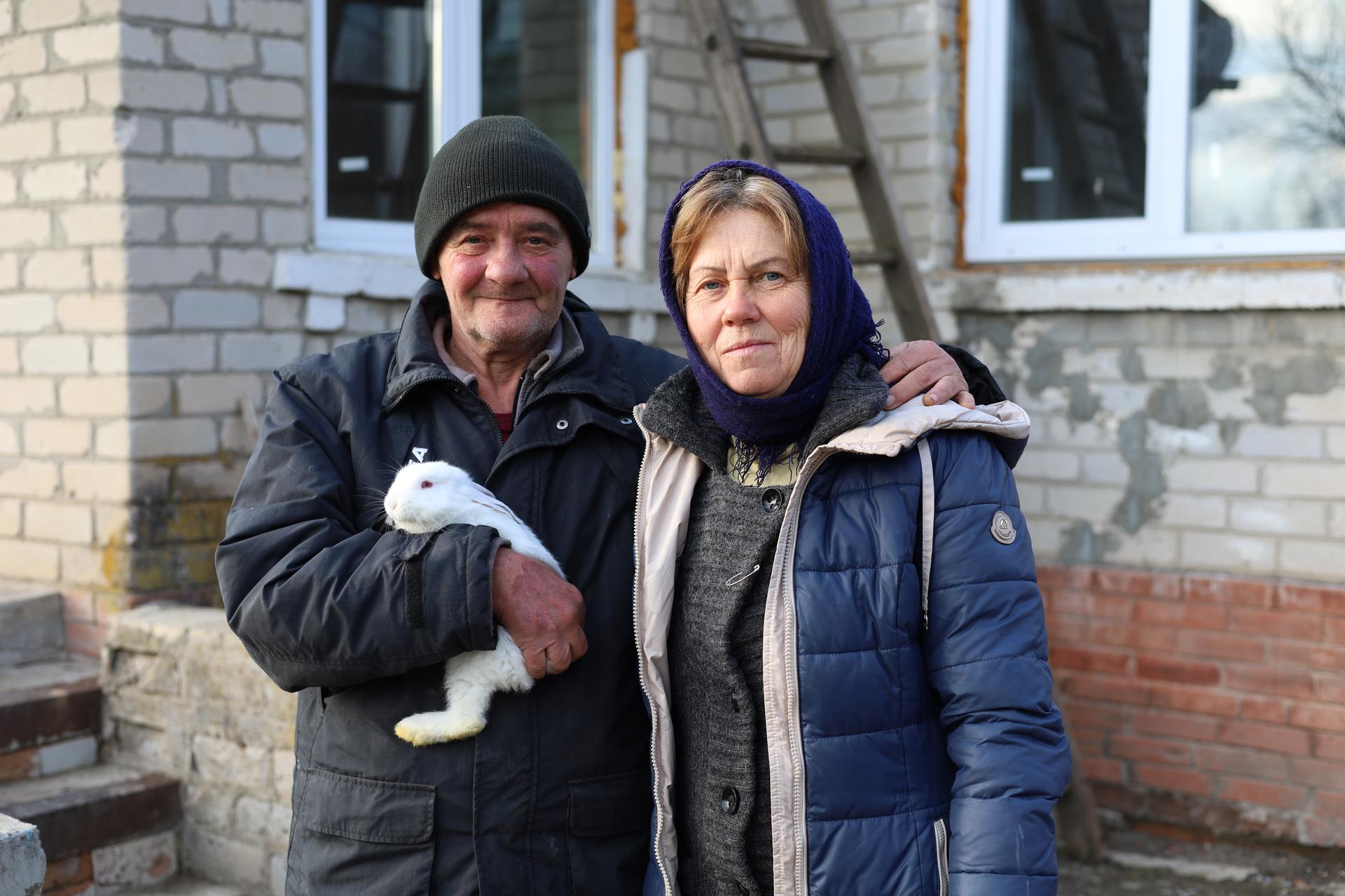 Ein älteres Ehepaar in dicken Winterjacken in einem Dorf in der Ukraine. Er hat ein weißes Kaninchen auf dem Arm.